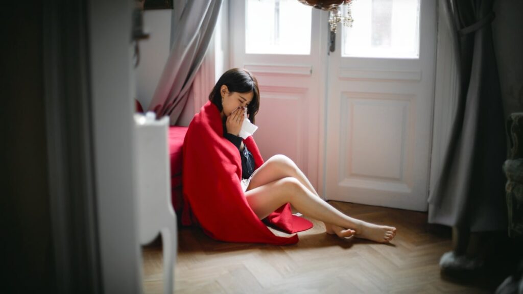 明るい部屋の床に赤いブランケットをかけて座る人物の写真。部屋には白いドアと窓、シャンデリア、グレーのカーテンがある。