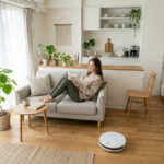 自然光が入るナチュラルな一人暮らしのワンルームで、ソファに座って読書をする女性。観葉植物やロボット掃除機がある、清潔感のあるミニマルな部屋。