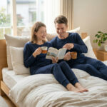 明るい寝室のベッドで、ネイビーのリカバリーウェアを着た男女が飲み物を片手に読書をしながらくつろいでいる