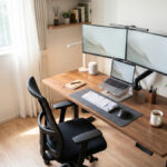 自然光が入る明るいホームオフィス。木目の昇降デスクにデュアルモニター、ノートPC、エルゴノミクスチェアを配置した快適なリモートワーク環境。