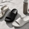 コンクリート床に並べられた白と黒の軽量サンダル、タオル、ボトル。ジムやプール、室内履きにも使いやすいリカバリーサンダルのイメージ