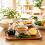 木のテーブルに並んだご飯、焼き鮭、野菜、レモン、調味料を使った健康的な和食の食卓
